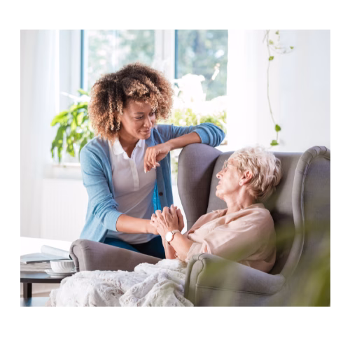 A lady is sat in an armchair turned towards a female carer deep in conversation clasping her hand.